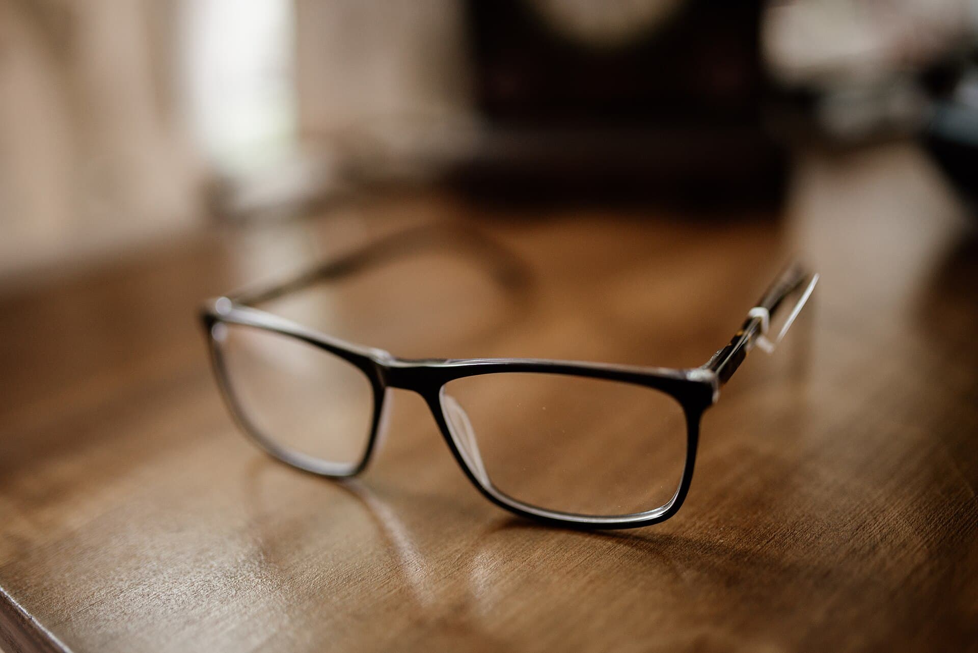 Black eyeglasses resting on a wooden desk in daylight, suggesting careful reading of language